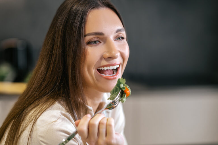 Uśmiechnięta kobieta z długimi brązowymi włosami trzyma widelec z sałatką, zbliżając go do ust.